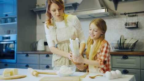 Beautiful-young-mother-preparing-everything-for-cooking,-pouring-the-flour-in-the-bowl-and-her-daughter-helping-her.-Nice-modern-kitchen.-Indoor